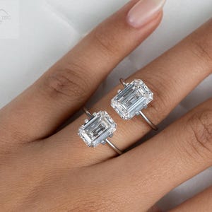May include: Two emerald cut diamond engagement rings on a hand. The rings are set in a simple band and feature a large, clear diamond.