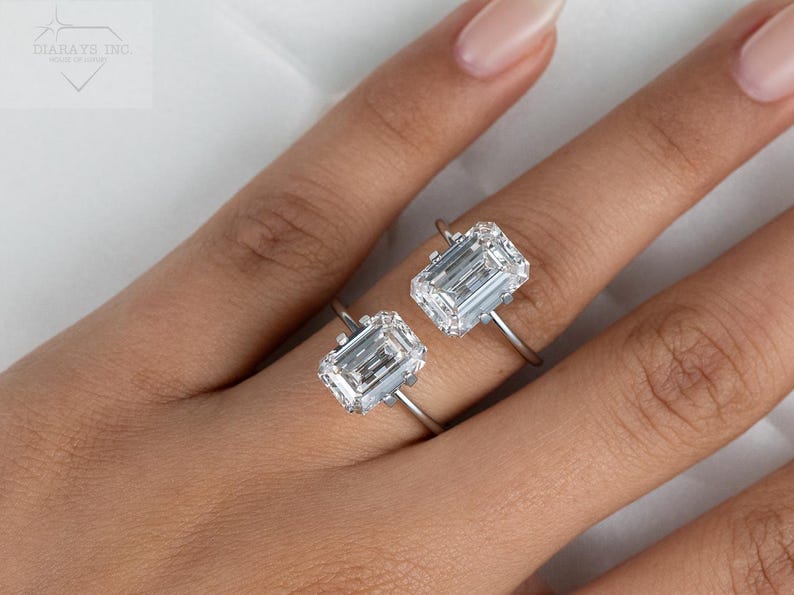 May include: Two emerald cut diamond engagement rings on a hand. The rings are set in a simple band and feature a large, clear diamond.