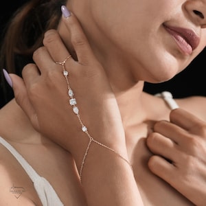 May include: A rose gold hand chain with multiple clear, sparkling gemstones. The chain connects to a ring and drapes across the back of the hand, with a delicate chain that attaches to the wrist. The gemstones are various shapes and sizes.