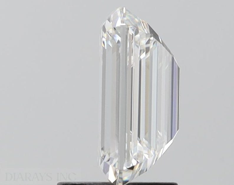 May include: A clear, rectangular emerald cut diamond with a brilliant finish. The diamond is set in a prong setting and is on a white background.