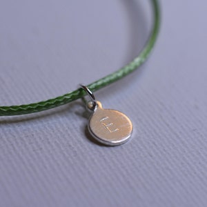 May include: A green cord bracelet with a silver charm that has the letter "E" engraved on it.