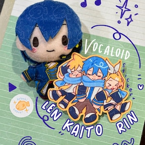 May include: A Vocaloid sticker featuring the characters Len, Kaito, and Rin. The sticker is a light blue, yellow, and white design with the characters' names written below. The sticker is on a green and white lined notebook with a blue plush toy of Kaito in the background.