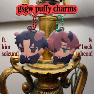 Puede incluir: Dos dijes inflados con personajes estilo anime, uno de cabello oscuro y ojos rojos, otro de cabello castaño y ojos verdes, ambos con traje. Texto: gsgw puffy charms ft. kim soleum! & baek saheon! con cadenas de bolas rojas y verdes.