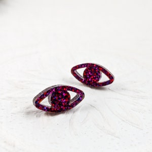 Könnte beinhalten: Ein Paar augenförmige Ohrringe mit glitzerndem rot-violettem Finish.