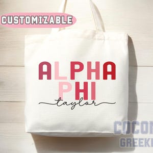 May include: White tote bag with the text "ALPHA PHI taylor" in shades of pink and red. The word "CUSTOMIZABLE" is above the text. The bag has a simple design and is likely used for carrying items.