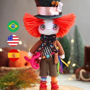 Peut inclure: Une poupée en crochet du Chapelier Fou d'Alice au pays des merveilles. La poupée porte un costume marron, une écharpe colorée et un grand chapeau vert avec un ruban rose et une étiquette indiquant "10/6". La poupée a les cheveux roux, la peau blanche et de grands yeux verts.