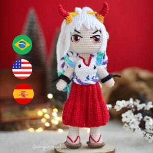 Pode incluir: Uma boneca de crochê com cabelo branco, chifres vermelhos e uma saia vermelha. A boneca está vestindo uma camisa branca com um padrão colorido e está em pé sobre uma base de madeira. A boneca tem uma expressão feroz no rosto.
