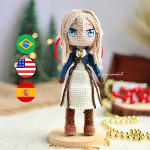 Pode incluir: Uma boneca de crochê com cabelo loiro, olhos azuis e um vestido branco. A boneca está em pé sobre uma base de madeira e está cercada por contas douradas. A boneca está usando botas marrons e uma jaqueta azul. A imagem também inclui três bandeiras: Brasil, Estados Unidos e Espanha.