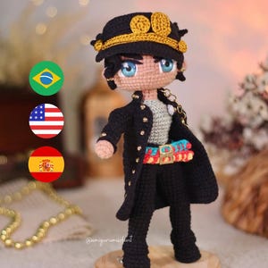 May include: Crocheted amigurumi figure of a character wearing a black jacket, black pants, and a gold hat with a gold chain. The figure is standing on a wooden base. The figure has blue eyes and a light skin tone.