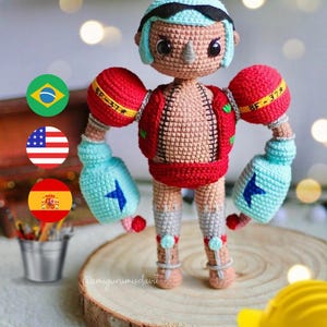 Op de afbeelding: Een gehaakt speelgoedfiguur van een bokser met een rode broek, blauwe handschoenen en een blauw hoofdband. Het speelgoed heeft een bruin lichaam en een kleine, zwarte neus. Het speelgoed staat op een houten oppervlak met een gele bouwhelm op de voorgrond.