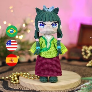 May include: A handmade crochet doll with green hair styled in pigtails and a matching green outfit. The doll has large blue eyes and is wearing a maroon skirt. The doll is standing on a wooden base.