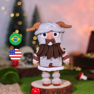 Puede incluir: Un guerrero de juguete de crochet con un casco gris, barba marrón y armadura blanca. El juguete está de pie sobre una rodaja de madera con un hongo rojo delante. El fondo es una imagen borrosa de un bosque.