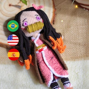 Pode incluir: Boneca de crochê com cabelo comprido e preto, vestindo um vestido estampado em rosa e branco, uma jaqueta marrom e uma máscara facial verde e marrom. A boneca tem olhos roxos e um laço rosa no cabelo.