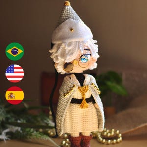 Op de afbeelding: Een handgemaakte gehaakte pop met wit haar, een puntmuts met een gouden stud en een ronde bril met een gouden montuur. Het figuur draagt een witte tuniek, een zwarte riem, een grijze cape met gouden accenten en bruine laarzen met blauwe ogen.