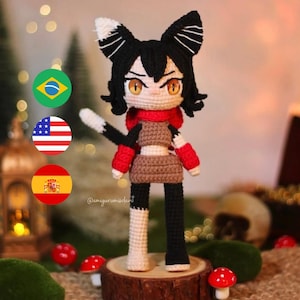 May include: A handmade crochet doll with black and white hair, cat ears, and orange eyes. The doll wears a red scarf, brown top, and black and white pants. It stands on a wooden slice with red mushroom decorations. The doll's creator's Instagram handle is visible.