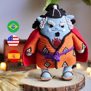 Może przedstawiać: Pluszowa zabawka amigurumi w postaci niebieskiego ogra, ubrana w pomarańczowo-czerwoną szatę z fioletowym pasem. Ogr ma czarny czubek głowy i białe włosy. Zabawka stoi na drewnianej platformie. @amigurumisa