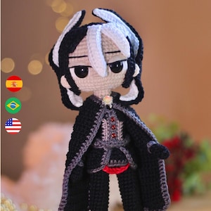 May include: A handcrafted crochet doll featuring a character with black and white hair, large black eyes, and a pale complexion. The figure wears a black cloak with grey trim, a dark jacket with three red buttons, and a small iridescent brooch.