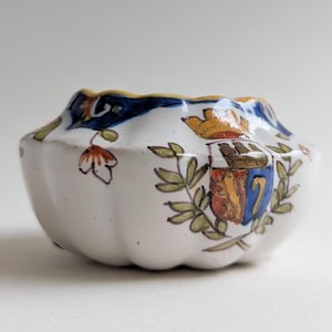May include: A small, white ceramic bowl with a scalloped rim. The bowl is decorated with a blue and gold floral pattern and a crest with a shield and crown. The bowl is approximately 3 inches in diameter.