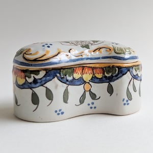May include: A ceramic trinket box with a lid. The box is white with blue, yellow, green, and orange floral and scrollwork designs. The lid has a curved shape and a blue border. The box has a scalloped edge.