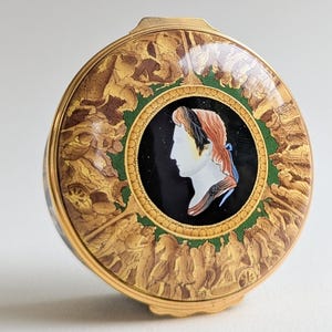 May include: A circular, gold-coloured compact featuring a profile portrait. The portrait is set against a black background, framed by a green border and a detailed gold-toned scene. The compact has a hinged lid and is highly decorative.