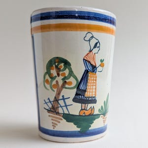 Könnte beinhalten: Ein Keramikbecher mit handgemaltem Design. Der Becher hat einen blau-orange gestreiften Rand und eine Szene einer Frau in traditioneller Kleidung, die Obst in der Nähe eines Baumes hält. Das Design ist vor einem weißen Hintergrund.