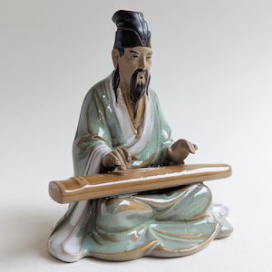 Op de afbeelding: Een keramieken beeldje van een zittende man die een snaarinstrument bespeelt. De figuur draagt een zwarte hoed en een lange jurk in groen- en wittinten. Het instrument is bruin en het beeldje heeft een serene uitdrukking.