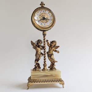 Peut inclure: Une horloge dorée antique avec un cadran rond et des chiffres romains. L'horloge est soutenue par un support décoratif avec deux chérubins et une base en marbre. Le design de l'horloge est orné et vintage.