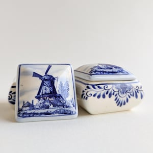 May include: Two ceramic trinket boxes with blue and white windmill and floral designs. The square box features a windmill scene, while the other is a small, lidded box with floral patterns. The boxes are on a white surface.