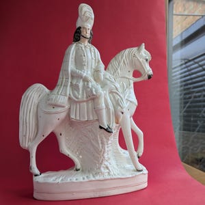 Puede incluir: Figurilla de cerámica blanca de una persona a caballo. La persona lleva un sombrero y una capa larga. El caballo está de pie sobre una base.