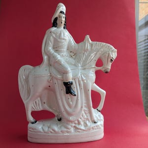 Op de afbeelding: Een wit keramiek beeldje van een persoon die op een paard rijdt. Het beeldje is in vintage stijl en heeft zwarte details op de kleding en het gezicht.