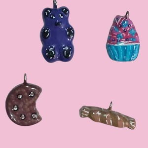 Könnte beinhalten: Vier handgefertigte Charms aus Polymer Clay: ein lila Gummibärchen, ein rosa-blauer Cupcake, ein brauner Schokoladenkeks und ein brauner und weißer Zuckerstangen.