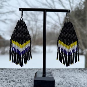 May include: A pair of black beaded earrings with yellow, white, and blue accents hanging from a black stand. The earrings are in the shape of a teardrop and have a fringe of black beads.