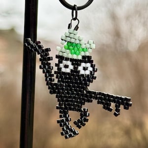 Può includere: Un ciondolo fatto a mano con perline a forma di creatura, con perline nere, bianche e verdi. Il ciondolo è sospeso a un anello di metallo nero. La creatura ha le braccia tese e un cappello verde.