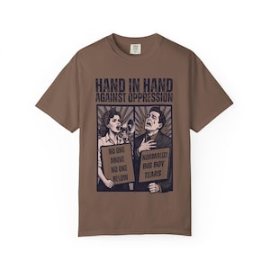 May include: A brown short-sleeve t-shirt featuring a graphic print with the text "HAND IN HAND AGAINST OPPRESSION". The illustration shows two people holding signs that read "NO ONE ABOVE NO ONE BELOW" and "NORMALIZE BIG BOY TEARS".