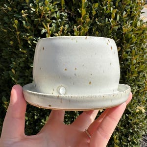 May include: A handmade, off-white ceramic planter with a matching saucer. The planter has a rounded shape with a slightly speckled texture and small brown spots. The saucer is round and fits snugly beneath the planter.