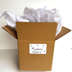 May include: A brown cardboard box filled with white tissue paper. A white rectangular label is attached to the front of the box with the handwritten black text "Mystery Pottery" surrounded by several question marks.