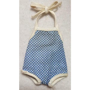 Op de afbeelding: Een blauw en wit polkadot poppenbadpak met crèmekleurige rand en haltertop. Het badpak heeft een strik aan de bovenkant en is gemaakt van een zachte stof. Het ontwerp is vintage en charmant.