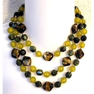 Puede incluir: Un collar de tres hilos con cuentas en tonos verdes, marrones y negros. El collar presenta una mezcla de cuentas redondas y geométricas, creando un efecto de capas. Las cuentas están ensartadas en un delicado cordón verde.
