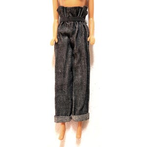 May include: Doll-sized denim pants with a high, ruffled waist and a black elastic belt. The pants are a dark gray color with a subtle textured pattern. The cuffs are rolled up at the ankles.