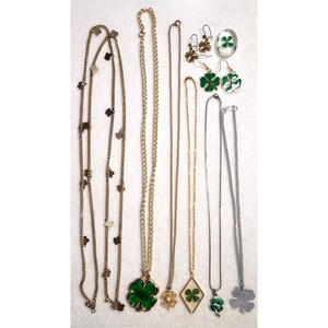 Op de afbeelding: Een collectie sieraden met klavertjevier-motieven, waaronder een lange goudkleurige ketting met klaveraccenten, diverse kettingen met hangers met groene en metalen klaverbedels, en twee paar oorbellen in de vorm van een klavertje.