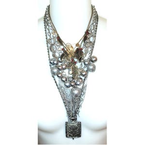 May include: A multi-strand statement necklace featuring silver-tone chains, metallic grey spherical beads, and intricate gold-tone leaf-shaped charms with small red accents. The design culminates in a square, textured pendant at the bottom centre.