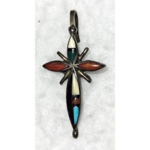 May include: A silver-toned cross pendant with inlaid gemstones. The cross features red, white, green, black, and turquoise stones. The pendant has a loop at the top for a chain and a small decorative bead at the bottom.