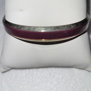 May include: A silver-toned bangle bracelet with a deep burgundy enamel center. The bracelet has a smooth, polished finish and a simple, elegant design. This piece of jewelry is suitable for everyday wear or special occasions.