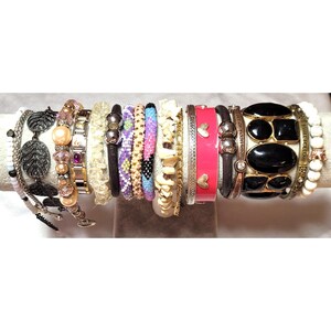 May include: A collection of various bracelets including a dark metal leaf design, peach-colored beads, a link bracelet with purple stones, clear crystal chips, beaded strands in purple and blue, white stone chips, a pink enamel cuff with hearts, and black stones.