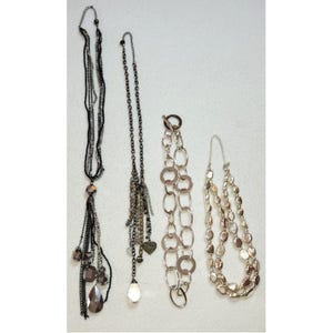 Puede incluir: Se muestran cuatro collares de varios estilos sobre un fondo blanco. Los collares incluyen un collar de borlas negro y plateado de múltiples hilos, un collar de cadena con un dije de corazón y un collar de cuentas en capas.