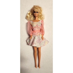 May include: A doll with blonde hair styled in a side-swept fringe, wearing a short dress featuring a pastel floral pattern in shades of purple, pink, and green. The dress has sheer, long, light pink sleeves and a fitted bodice with a pink waistband.
