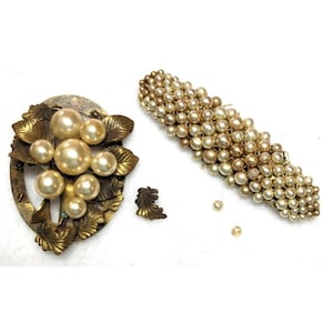 Puede incluir: Juego de joyas vintage con un broche dorado con perlas de imitación y detalles de hojas, una horquilla cubierta de perlas y un pequeño pendiente en forma de hoja. Las perlas son de color crema y el metal tiene un aspecto envejecido.