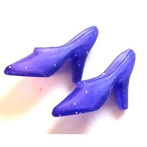 May include: A pair of miniature, translucent purple high heels with pointed toes and stiletto heels. The shoes have small white dots scattered across their surface, and are set against a white background.