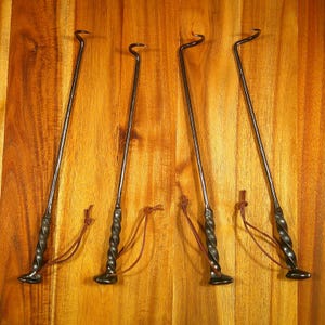 Puede incluir: Cuatro herramientas de metal negro para fogatas con mangos retorcidos y extremos en forma de gancho. Cada herramienta tiene una base peque&ntilde;a y plana y un lazo de cuero marr&oacute;n. Las herramientas est&aacute;n dispuestas sobre un fondo de madera.