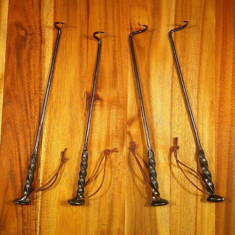 Puede incluir: Cuatro herramientas de metal negro para fogatas con mangos retorcidos y extremos en forma de gancho. Cada herramienta tiene una base peque&ntilde;a y plana y un lazo de cuero marr&oacute;n. Las herramientas est&aacute;n dispuestas sobre un fondo de madera.